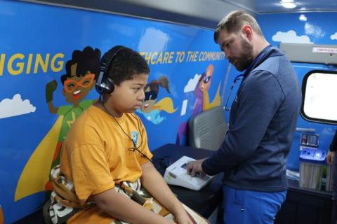 A young patient wearing headphones receives a hearing test from a healthcare provider inside the Pediatric Mobile Clinic, decorated with superhero-themed murals.