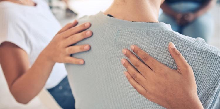 Supportive hands on the back of someone in group therapy, grief, sexual assault, intimate partner violence, rape, sexual violence