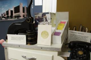 Display of historical hospital artifacts, including a seal press used by Minneapolis General Hospital until 1963, patient file cards, a black rotary telephone, and a gold-embossed certificate, all arranged on a nurse’s station exhibit.
