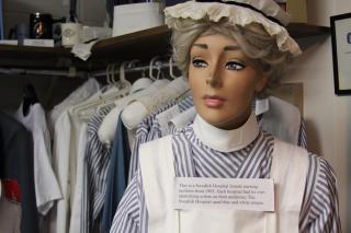 A mannequin dressed in a blue-and-white striped 1905 Swedish Hospital nursing uniform, displayed in a historical exhibit with clothing and accessories in the background. A sign explains the uniform’s origin and color significance.