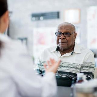 An older man wearing glasses and a striped shirt sits across from a healthcare professional in a clinical setting. The doctor, whose back is to the camera, gestures with their hands while the patient listens attentively.