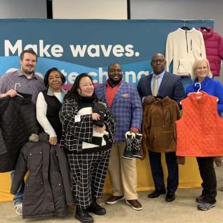 Group of six adults smiling and holding winter clothing donations in front of a sign that reads "Make waves."