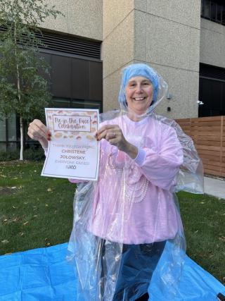 Woman smiling outdoors wearing a pink sweatshirt, blue hair cap, and clear plastic poncho, holding a sign that reads “Pie in the Face Celebration – Thank you for pieing Christene Jolowsky. Everyone raised $300.”