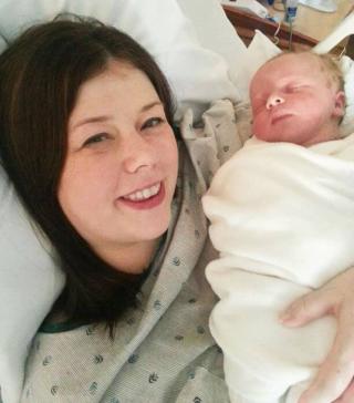 Smiling mother in a hospital bed holding her newborn baby wrapped in a white blanket.