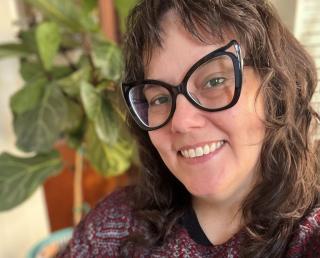 Smiling person with long dark hair and large black glasses, wearing a red patterned sweater, sitting indoors near a leafy plant.