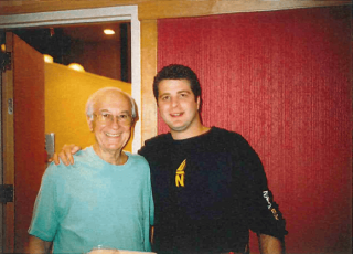 Two men smiling and posing together indoors; one older man wearing a light blue shirt and one younger man in a dark shirt with a yellow logo.