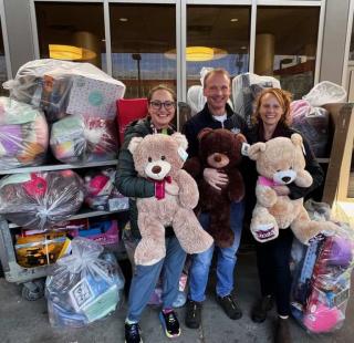Three smiling adults standing outside in front of large bags filled with toys, each holding a large teddy bear, as part of a toy donation event.