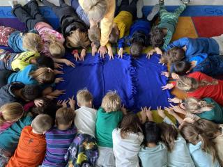 Group of young children and a teacher laying in a circle on a colorful classroom rug, reaching toward a blue fleece blanket with knotted edges placed in the center.