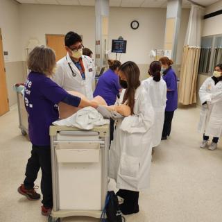 Students in lab coats practicing medical skills on a mannequin with instructor guidance.