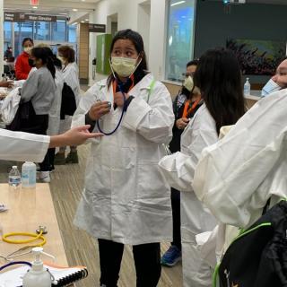 Students in lab coats practicing with medical equipment at a health education event.