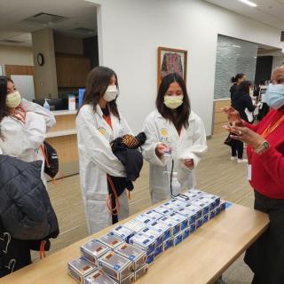 Students in lab coats receiving materials and talking to a staff member at a table.