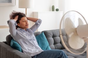 woman sitting on a couch cooling off with a fan, Keeping your cool when it's hot, heat stroke, hotter temperatures are associated with poorer sleep, drink liquids, Dr. Kristi White